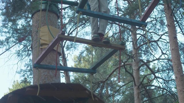 A teenager in a safety harness passes a dangerous section of a high rope course in a rope town. A guy balances on suspended wooden frames against the background of tree crowns on a summer day.