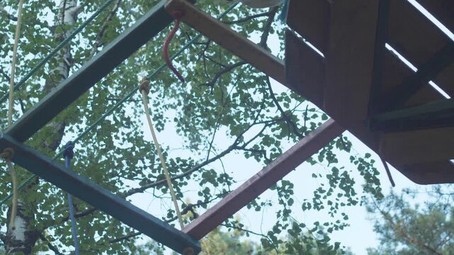 A teenager in a safety harness successfully completes a high rope course stage and steps onto the platform. A guy overcomes an obstacle of suspended wooden frames in a summer rope park.