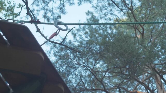 The hands of a teenager re-attach a safety ring onto a metal cable in a rope park. The equipment element smoothly slides along the stretched line against the background of tall pine trees