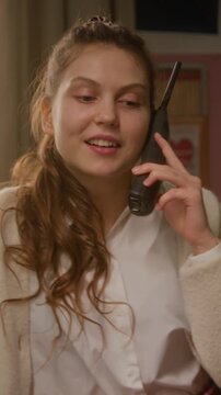 Vertical video of flirty Caucasian teenager talking on vintage cordless phone, smiling and twirling hair. Ideal for nostalgic brand advertisements or retro-themed social media content