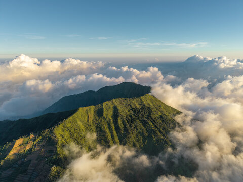 Aerial view of Mount Bismo peak rising above a sea of clouds with lush green slopes and terraced agricultural fields in Kecamatan Kejajar, Jawa Tengah, Indonesia.