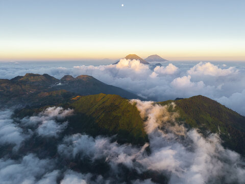Aerial view of the lush green slopes of Gunung Bismo rising above a sea of clouds at sunrise in Kecamatan Kejajar, Jawa Tengah, Indonesia.