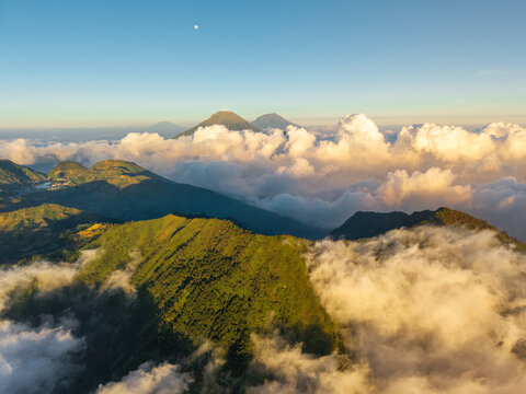 Aerial view of the lush green slopes of Mount Bismo near the Sikunang basecamp, surrounded by clouds under a clear sky at sunset in Kecamatan Kejajar, Jawa Tengah, Indonesia.
