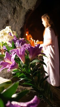 Cave shrine with lilies and candles, woman in silent devotion.
