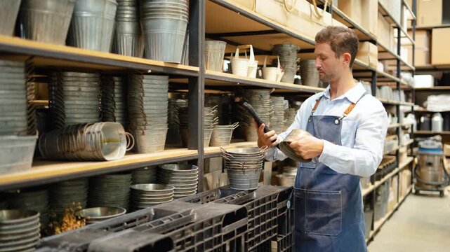 Male small business owner wearing a denim apron uses a handheld barcode scanner to check inventory of galvanized metal planters on warehouse shelves. Retail stock management logistics concept.