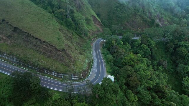 Serpentine mountain road cutting through misty tropical forest and rugged green hills beneath drifting low clouds, pullback aerial drone footage.