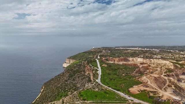 At Malta's Dingli Cliffs, the rugged sea cliffs and rocky terrain meet surrounding farmland and vegetation along the Mediterranean coastline under a cloudy sky in an aerial hyperlapse view.