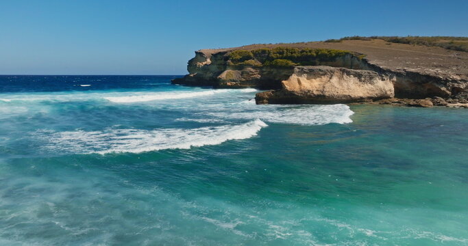 Ocean waves breaking on a rugged shore with a towering cliff and arid landscape, showcasing the power of nature and the beauty of the vast sea under a clear blue sky. Drone flight zoom out panorama