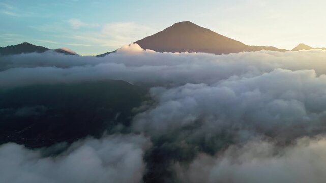 Cloud inversion surrounding a towering volcanic mountain illuminated by golden sunrise light above a sea of clouds, aerial drone footage.