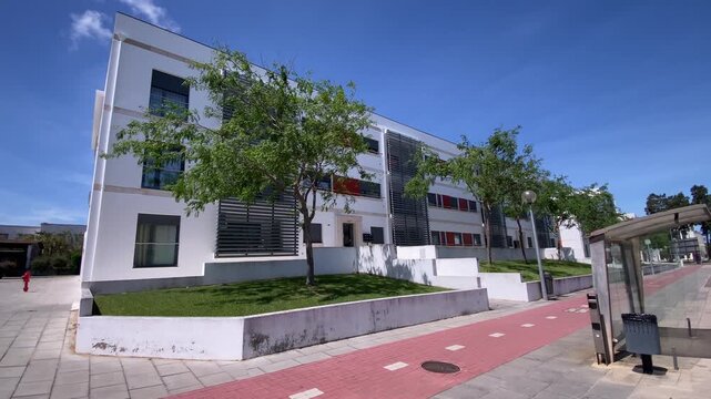 Modern architecture building facade with white walls and dark windows, featuring trees and a paved pedestrian path on a sunny day