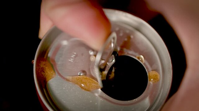 A close-up of a finger lifting the tab of a metal can to open it with condensation visible on the surface.