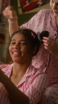 Vertical video of Caucasian teenager styling hair of African American girl friend during pajama party. Footage represents diversity and friendship for beauty or lifestyle brand commercials