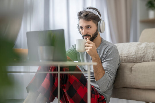 Man with headphones working on laptop at home while drinking coffee and wearing plaid pajama pants