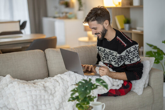 Caucasian European man in Christmas sweater using laptop and credit card on cozy couch at home