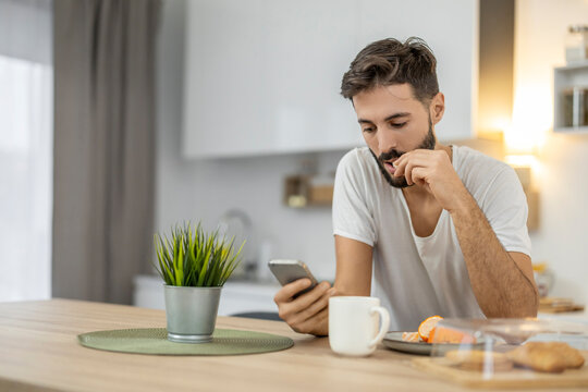 Young man checking smartphone while eating breakfast at home kitchen counter in casual morning routine