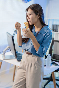 Asian businesswoman eating instant cup noodles while working at her office desk at night.