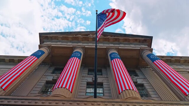 United States flag above neoclassical building columns, fabric banners cover four columns below clouds.
