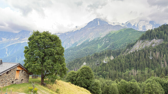 A rustic stone cabin overlooks a lush valley where a chairlift traverses dense pine forests, set against a backdrop of majestic snow-capped peaks under a dramatic, cloud-filled sky.