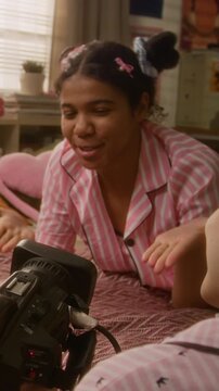 Vertical video of African American teenage girl in pajamas vlogging with friend on bed, gesturing and talking to camera. Footage is ideal for social media marketing and youth lifestyle campaigns