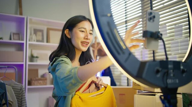 Young Asian woman fashion entrepreneur live streaming selling clothes online with smartphone ring light and laptop in home studio, representing small business owner and professional women.