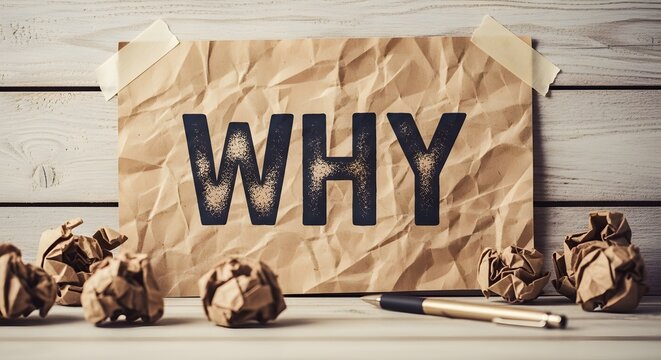 Crumpled paper note with the word why written on it sits on a wooden surface beside a pen.