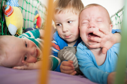 Big brother embraces his wailing baby sibling as another curious little one smiles inside their vibrant playpen