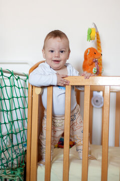 Joyful infant's bright smile shines while confidently standing in his cozy wooden crib, surrounded by soft nursery treasures