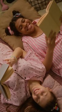 Vertical top view of Caucasian and African American teenage girls lying on bed in pajamas, reading books and laughing. Footage is perfect for youth lifestyle and education marketing campaigns