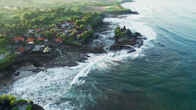 Hindu sea temple on coastal rocks surrounded by relentless ocean waves and white surf along a dramatic tropical shoreline showcasing one of Bali most iconic spiritual landmarks, aerial drone view