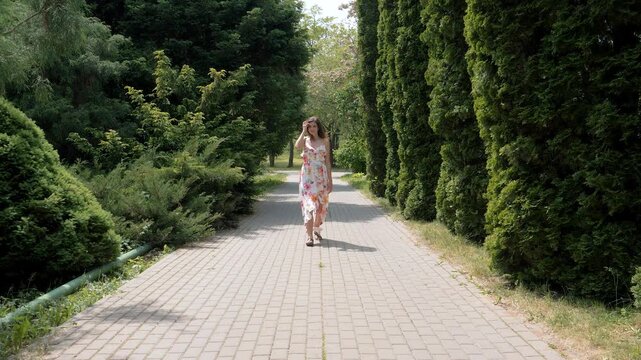 Pretty young woman wearing long floral dress walking forward on cobblestone path in green summer park surrounded by lush trees. Female enjoying the sunny weather and her leisurely stroll