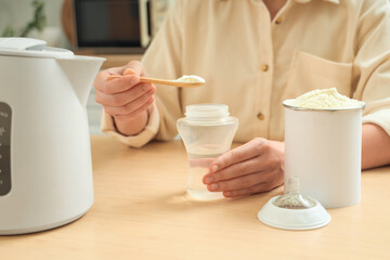 Woman with kettle preparing milk baby formula on table in kitchen, closeup