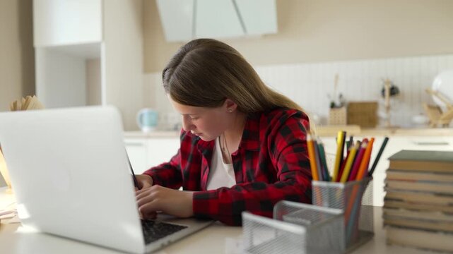 Teenage girl studying at home using laptop. Young student types on keyboard doing homework. Schoolgirl learns online at desk with lamp. Focused teen works on distance education task indoors.