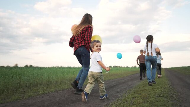 Children running with balloons through grass field at sunset. Kids run together holding colorful balloon strings. Happy children play in green meadow. Group of kids running outdoors in summer field.