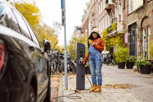 Smiling woman using smart phone and charging electric car at station