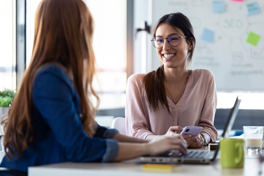 Smiling businesswomen using smartphone, talking with her colleague