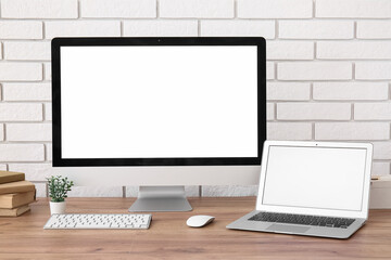 Fototapeta premium Modern computer monitor, laptop, keyboard, mouse and books on wooden table near light brick wall