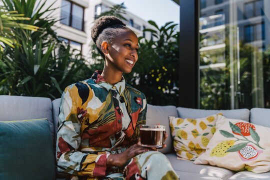 Portrait of happy young woman sitting on couch in garden drinking black coffee