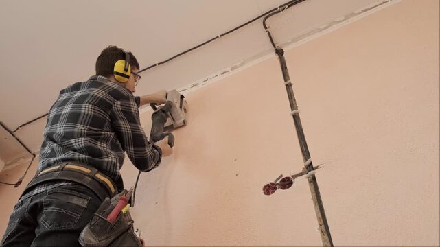 Electrician in plaid shirt safety glasses and ear protection cuts wall channels for electrical wiring installation work
