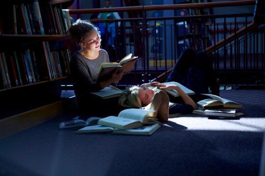 Female student lying on floor in a library with friend reaing book