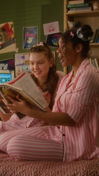 Caucasian and African American teenage girls sitting on bed in pajamas, reading magazine and laughing together. Clip is ideal for youth lifestyle and friendship marketing, vertical