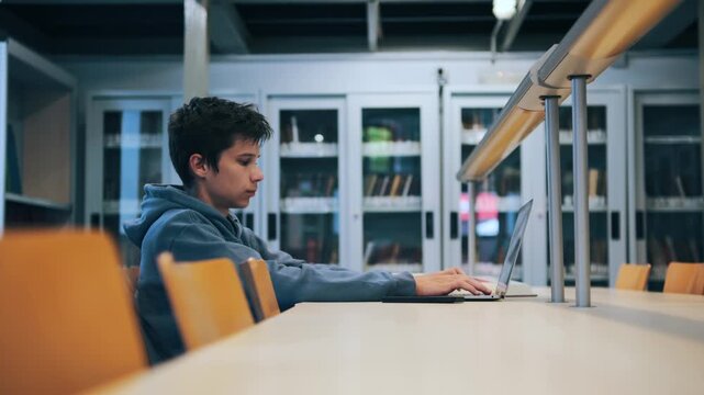 A focused male student sits at a desk in a university library, diligently working and typing on his laptop computer while preparing for exams.