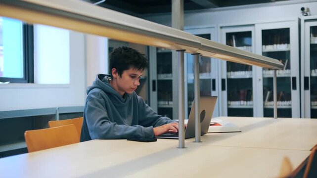 A focused teenage boy sits at a desk in a university library or study hall, diligently working and typing on his laptop computer for his studies.