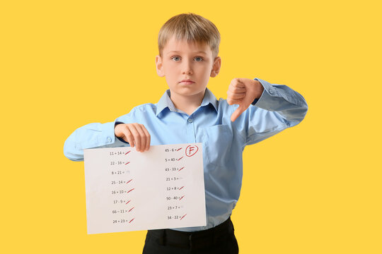 Sad schoolboy holding paper with bad results of school test and showing thumb-down gesture on yellow background