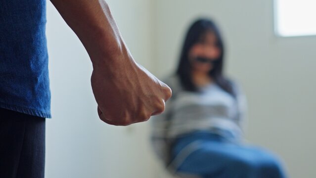 Criminal Clenches His Fist In The Foreground While A Scared Young Female Hostage Sits Gagged