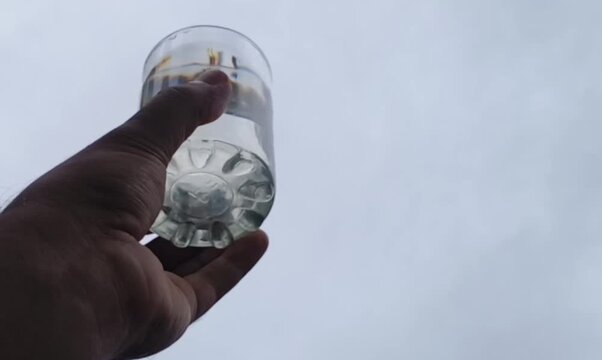 Drinking water glass or cup inside hand holding against the sky background in hot summer weather. Closeup of person checking clear natural fresh water for impurity.
