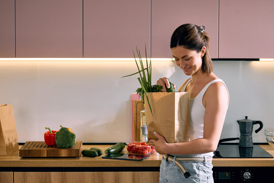Woman unpacking grocery bag while organizing fresh vegetables in modern kitchen. Concept of food preparation, healthy lifestyle, grocery delivery, sustainable shopping and meal organization.