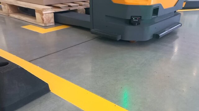 A warehouse electric forklift with long grey metal forks drives into a wooden pallet carrying cardboard boxes, positioned on a concrete floor with yellow safety markings during indoor operation.