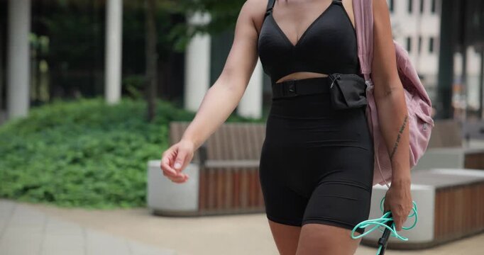 A young beautiful female athlete in sportswear with a backpack and a skipping rope goes for a workout in an urban park among high-rise buildings