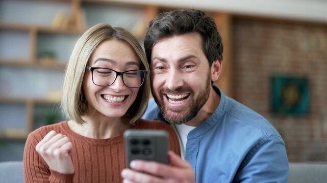 Happy excited married couple celebrating success reading great news on mobile phone sitting on sofa in living room at home. Smiling joyful husband wife satisfied with positive good message. Close up