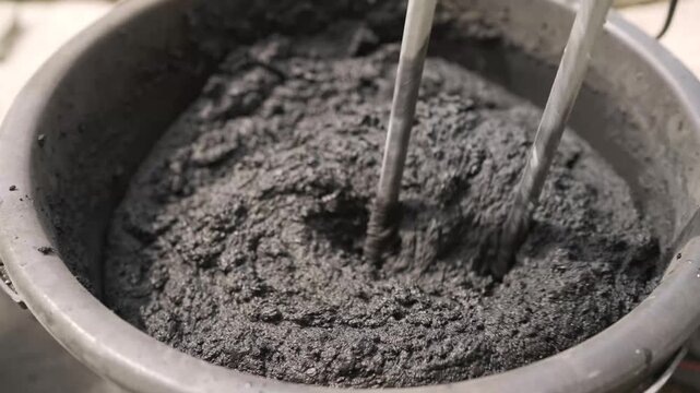 Close up of wet gray concrete being mixed in black plastic bucket using metal rods. Dark tones dominate with glossy wet texture